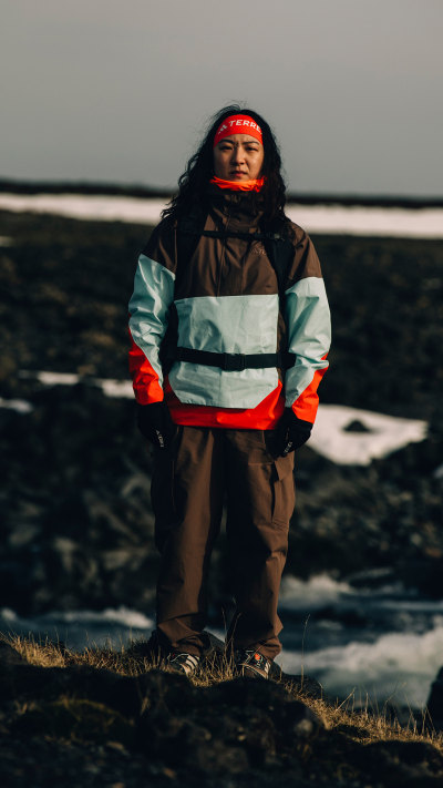 A person wearing a warm, colorful jacket stands on a grassy field with a body of water and a cloudy sky in the background.