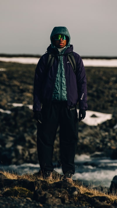 A person in a warm, hooded jacket stands on a grassy, rocky shore overlooking a body of water under a cloudy, dimly lit sky.