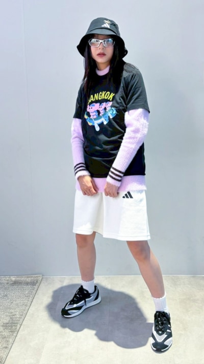 A person wearing a black t-shirt with a graphic design, a bucket hat, and white shorts stands on a concrete surface against a plain background.