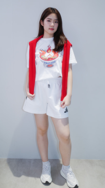 A young woman with long dark hair is wearing a red vest and a white t-shirt with a graphic design, standing against a plain white background.