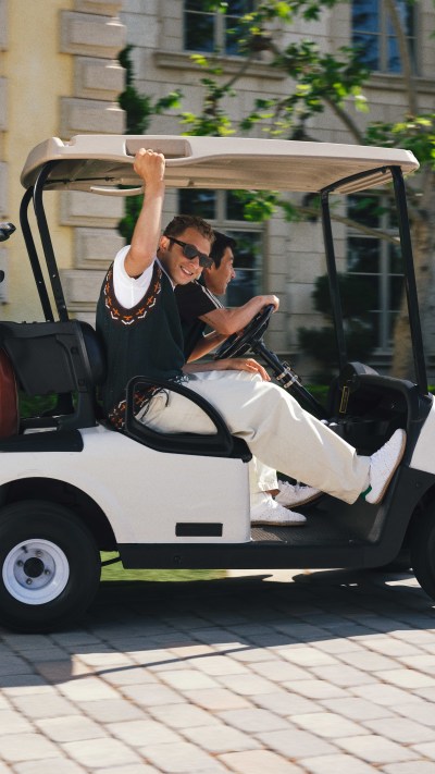 The image shows a person sitting in a white golf cart, with a building and greenery visible in the background.