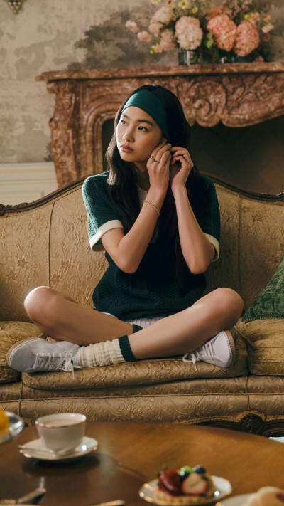 A young woman in a green dress sits cross-legged on a couch, surrounded by a floral arrangement and a cozy, ornate interior setting.
