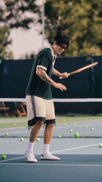 A person in a green shirt and white shorts is playing tennis on a tennis court surrounded by trees and greenery.