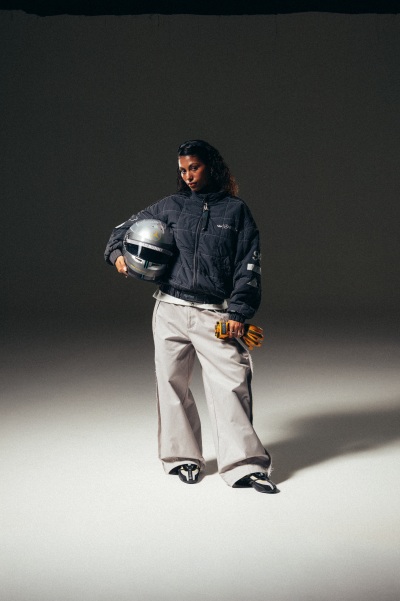 A person wearing a dark jacket and light-colored pants stands in a dimly lit environment, holding a helmet in their hands.