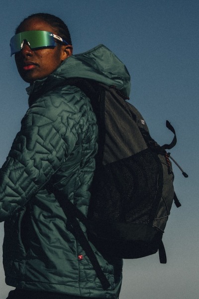 Osoba w zielonej, pikowanej kurtce i plecaku z okularami przeciwsłonecznymi stoi na tle błękitnego nieba.