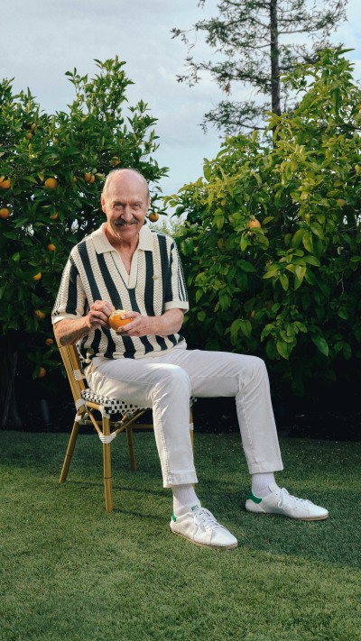 The image shows an elderly man sitting in a chair in a lush, green garden setting, wearing a striped shirt and white pants.