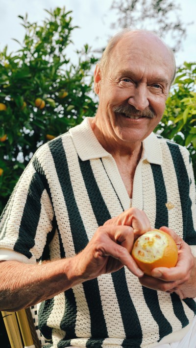 A smiling elderly man in a striped sweater holds a freshly picked apple in a lush, green garden setting.