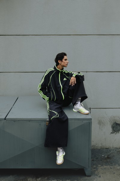 A person wearing a black and green tracksuit is sitting on a concrete structure against a plain gray wall.