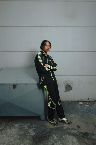 A person wearing a black and neon green athletic outfit stands in front of a plain gray wall.