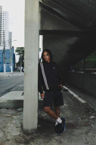 A person wearing a black jacket and shorts is standing under a concrete structure, with tall buildings visible in the background.