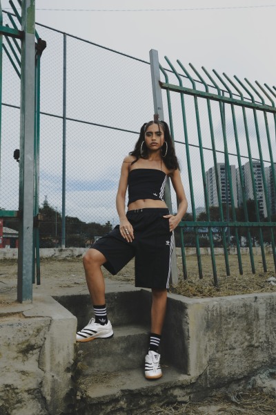 A young woman in a black crop top and shorts is sitting on a concrete structure in front of a metal fence, with buildings visible in the background.