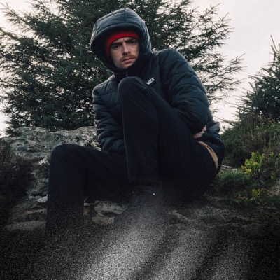 Človek v čiernej bunde s kapucňou a klobúkom sedí na prašnej ceste obklopenej stromami v zalesnenej oblasti.