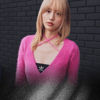 Una giovane donna dai capelli biondi che indossa un maglione rosa acceso e peloso sta di fronte a un muro di mattoni scuri.