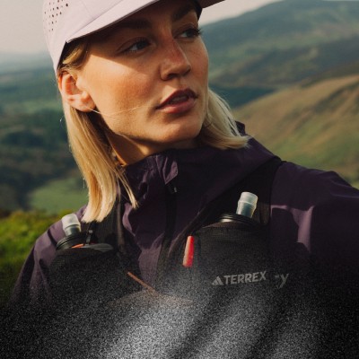 Una giovane donna che indossa una giacca Terrex viola e nera si trova in un paesaggio montuoso, con lo sguardo rivolto verso l'obiettivo.