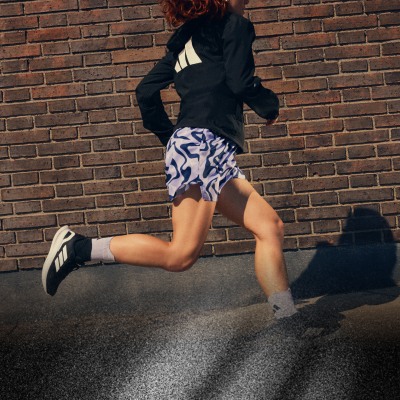 Človek v čiernej bunde a vzorovaných šortkách behá na pozadí tehlovej steny.