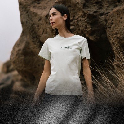 Una giovane donna che indossa una T-shirt bianca sta in un paesaggio roccioso e accidentato con un cielo nuvoloso sullo sfondo.