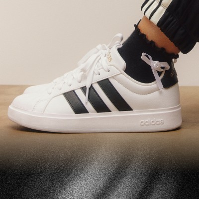 Un paio di sneaker adidas bianche e nere con lacci, poggiate su un pavimento di legno.