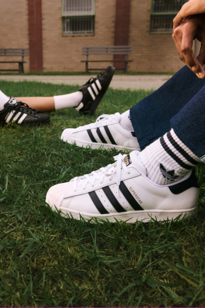 La imagen muestra un par de zapatillas adidas blancas y negras sobre un campo de hierba, con un edificio de ladrillo visible al fondo.