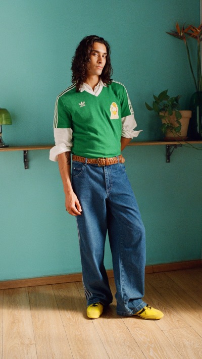 A person wearing a green jersey, blue jeans, and yellow shoes stands in a room with a green wall and shelves in the background.