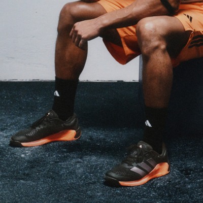 The image shows a person wearing orange athletic clothing and black and orange sneakers, sitting on a dark surface with a body of water in the background.