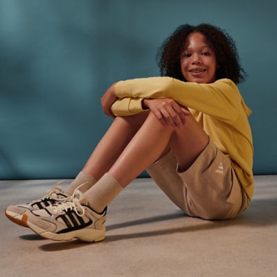 A young woman with curly hair sits on the floor, wearing a yellow top and beige pants, with sneakers on her feet, against a blurred teal background.