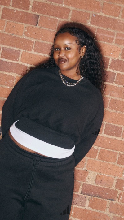 A smiling woman with curly hair stands in front of a brick wall, wearing a black outfit with a white stripe.