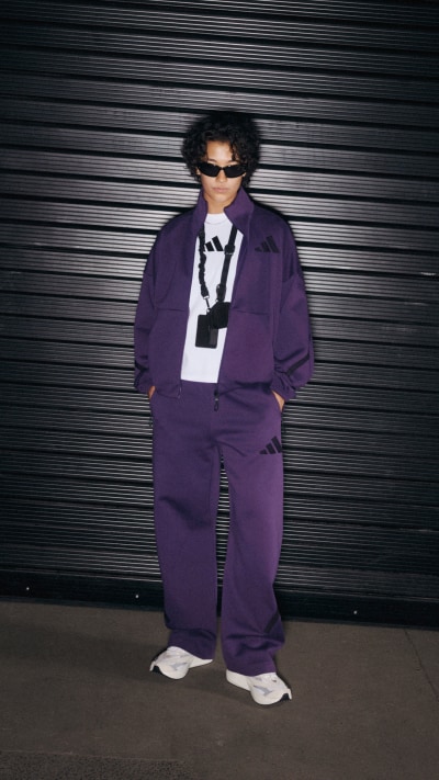 A person wearing a purple outfit, including a jacket and pants, stands in front of a dark, striped background.
