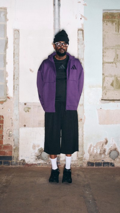 A person wearing a purple jacket and black pants stands in front of a dilapidated brick building.