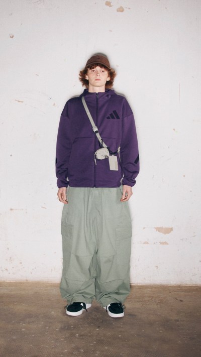 A person wearing a purple jacket and gray pants stands in a plain, white-walled room.