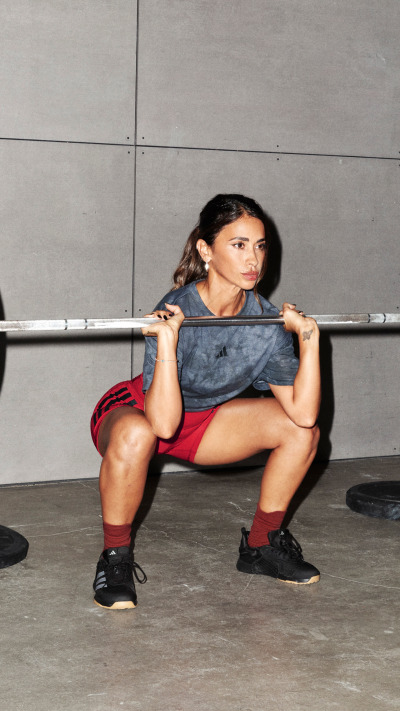 A woman in athletic clothing is performing a squat exercise while holding a barbell in a gym setting with a concrete wall in the background.