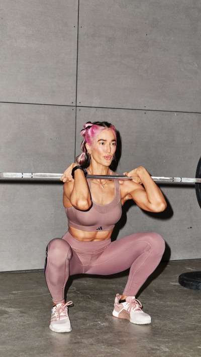 A woman with pink hair is performing a squat exercise while holding a barbell in a gym setting with a concrete wall in the background.