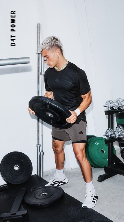 Eine muskulöse Person in einem schwarzen Hemd und Shorts sitzt auf einer Hantelbank, die von verschiedenen Trainingsgeräten in einem Fitnessstudio umgeben ist.