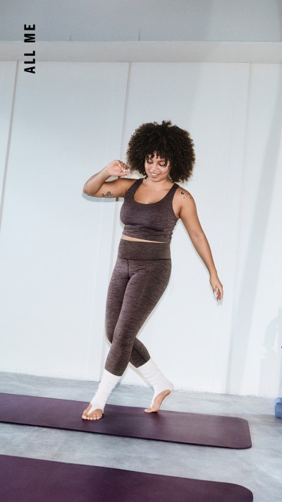 Eine Frau mit lockigem Haar steht in einem hellen, minimalistischen Raum auf einer Yogamatte.