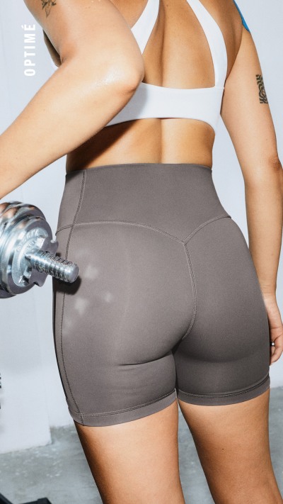 Das Bild zeigt eine Person im unteren Rumpfbereich in eng anliegenden grauen sportlichen Shorts, die eine Hantel in den Händen hält, vor einem schlichten weißen Hintergrund.