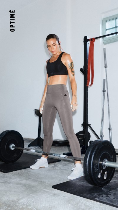 Eine Person, die einen schwarzen Sport-BH und graue Leggings trägt, steht in einem Fitnessstudio und im Hintergrund sind Trainingsausrüstung wie eine Langhantel und Hantelscheiben zu sehen.