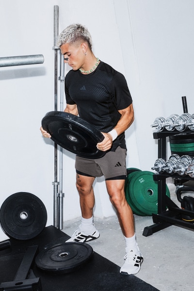 Eine Person in einem schwarzen Hemd und schwarzen Shorts hebt eine schwere Langhantel in einem Fitnessstudio, das von verschiedenen Trainingsgeräten umgeben ist.