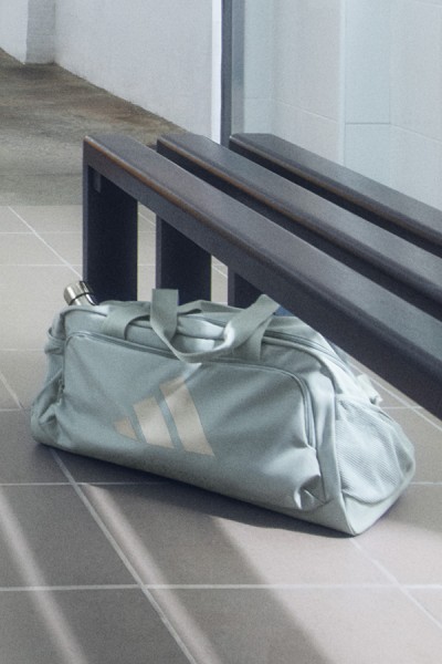 Eine graue Sporttasche sitzt auf einem Fliesenboden vor einer Holzbank vor einer weißen Wand.