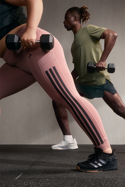 Two individuals, one in the foreground wearing pink athletic leggings and the other in the background holding dumbbells, are engaged in a fitness activity in a dimly lit setting.