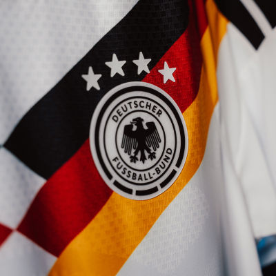 The image shows the logo of the German national football team, featuring a black eagle emblem on a white background with the German flag colors of black, red, and gold in the background.