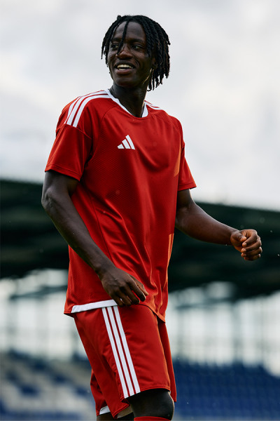 A smiling individual wearing a red Adidas sports jersey stands in front of a body of water and a cloudy sky.
