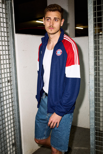 A young man wearing a blue and red jacket with the Bayern Munich logo stands in a hallway with metal grates in the background.
