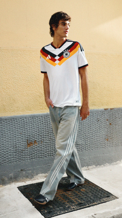 The image shows a young man wearing a white jersey with colorful geometric patterns and gray sweatpants, standing on a textured surface against a plain wall.