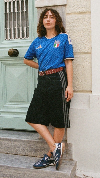 A person wearing a blue sports jersey and black shorts is standing on the steps of a building with arched windows and a wooden door in the background.