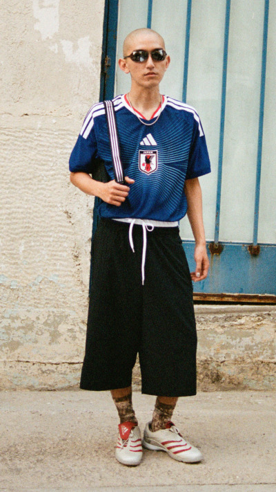 A person wearing a blue and white sports jersey, black shorts, and red and white sneakers stands in front of a weathered, blue-painted wall.