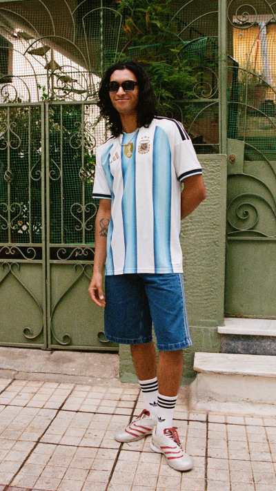 A person wearing a light blue and white jersey, blue shorts, and white sneakers stands in front of a decorative metal gate with a lush, green background.