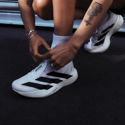 The image shows a person's lower body and feet, with the person wearing white sneakers and a tattoo visible on their arm.