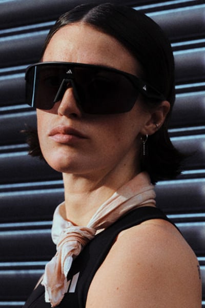 A person wearing sunglasses and a scarf stands in front of a metal shutter backdrop.