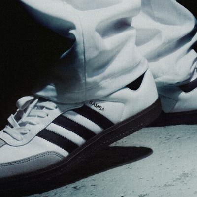 The image shows a pair of white and black Adidas Samba sneakers on a concrete surface.
