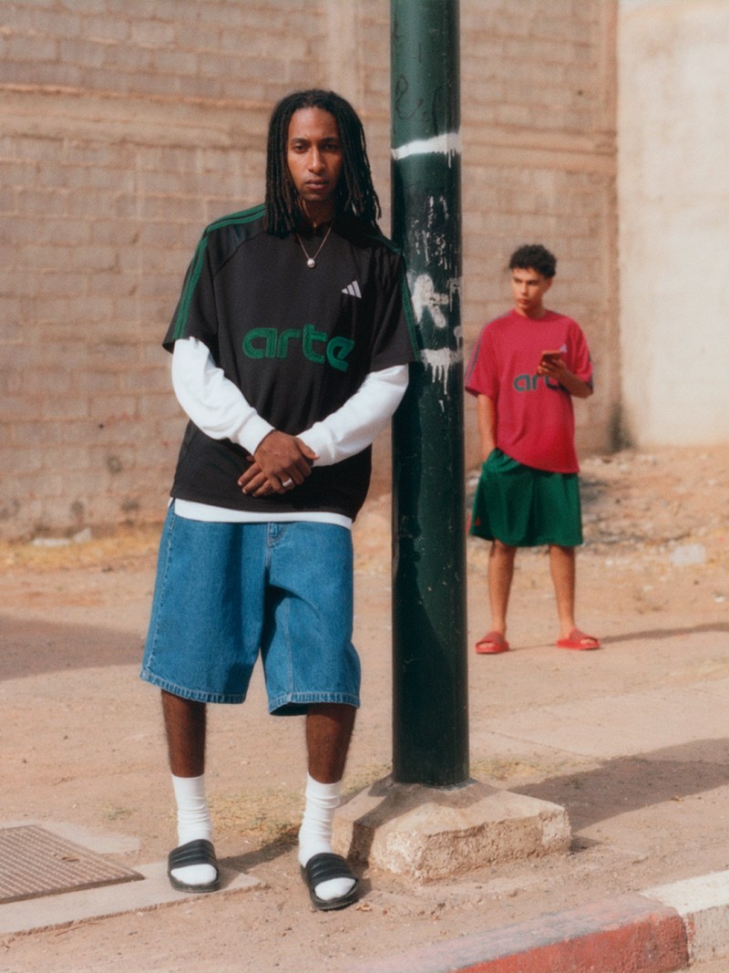 A man wearing a black and green shirt and blue shorts stands in the foreground, while a young boy in a red shirt is visible in the background against a brick wall.