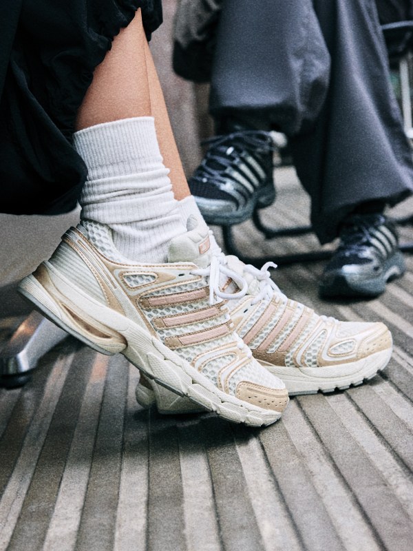 The image shows a pair of white and beige athletic shoes with intricate patterns and laces, resting on a wooden surface with other people's feet visible in the background.